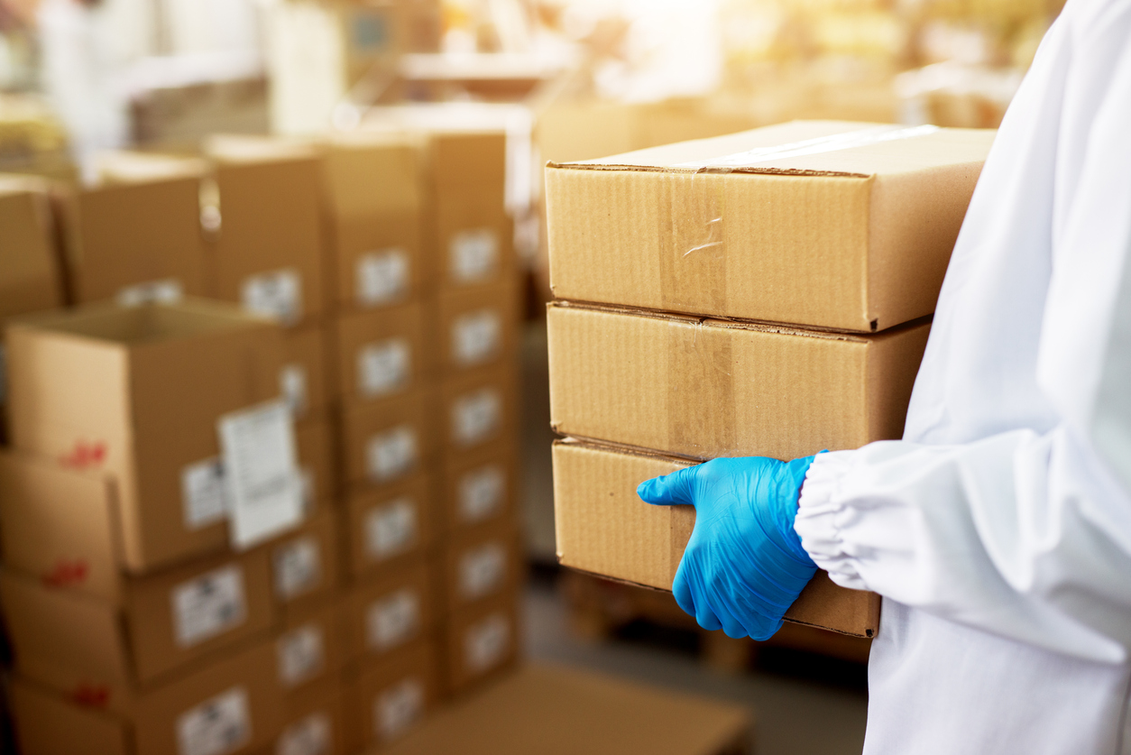 worker carrying a stack of duck taped brown boxes in factory storage room while wearing sterile cloths and rubber gloves.